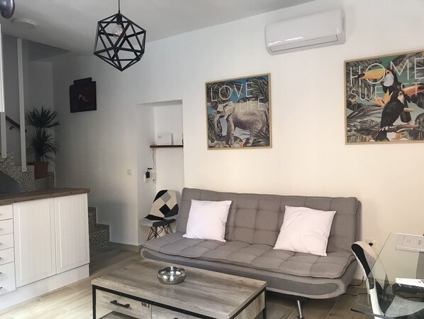 A modern living room with a grey sofa, a wooden coffee table, and a geometric pendant light. There are two wall-mounted paintings, one with a cow and the other with birds. A small shelf with a plant is visible in the background.