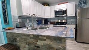 A kitchen with white cabinets, a stainless steel refrigerator, a microwave, and a stone island with a sink.