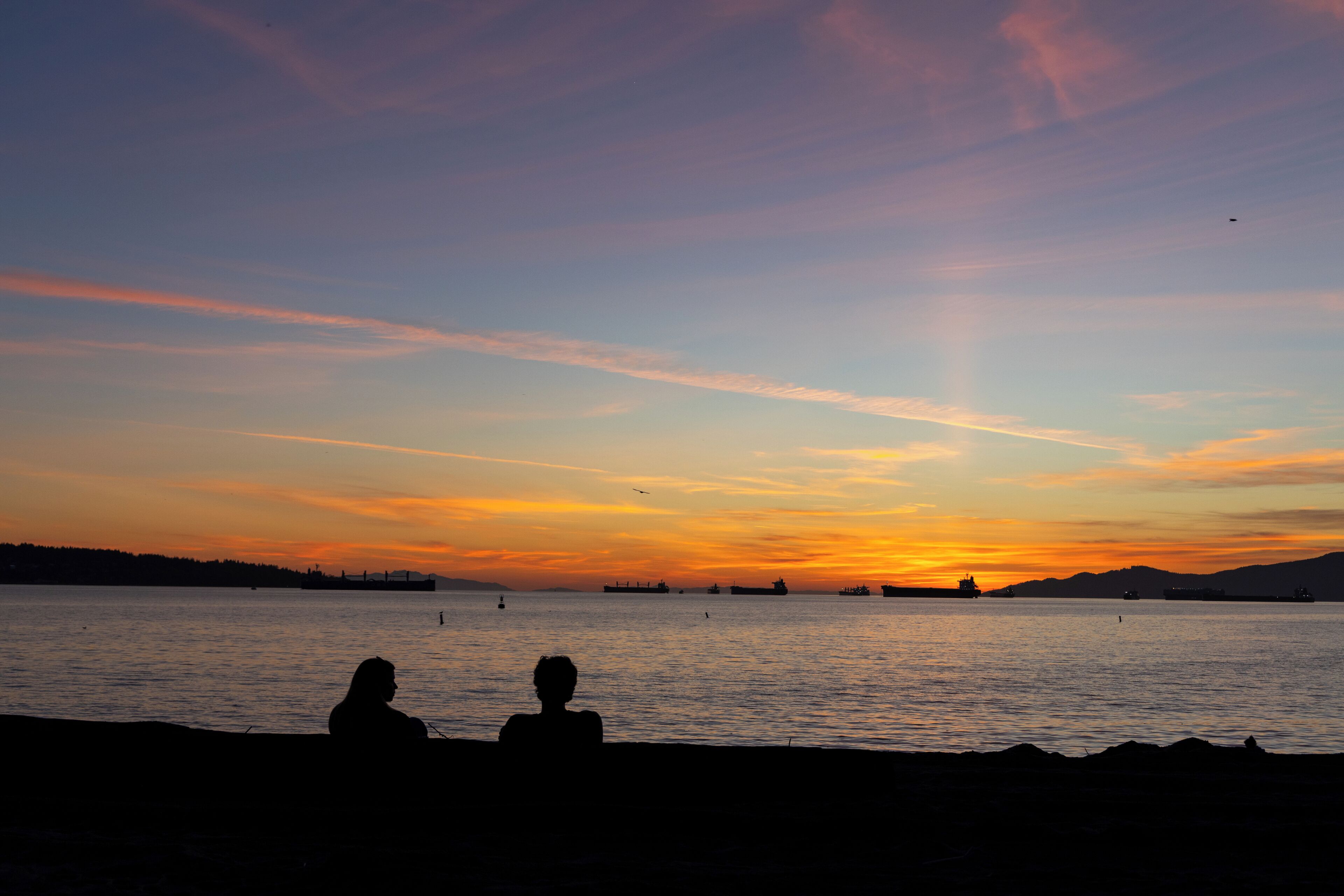 Silhuetas de duas pessoas sentadas à beira da água durante o pôr do sol.