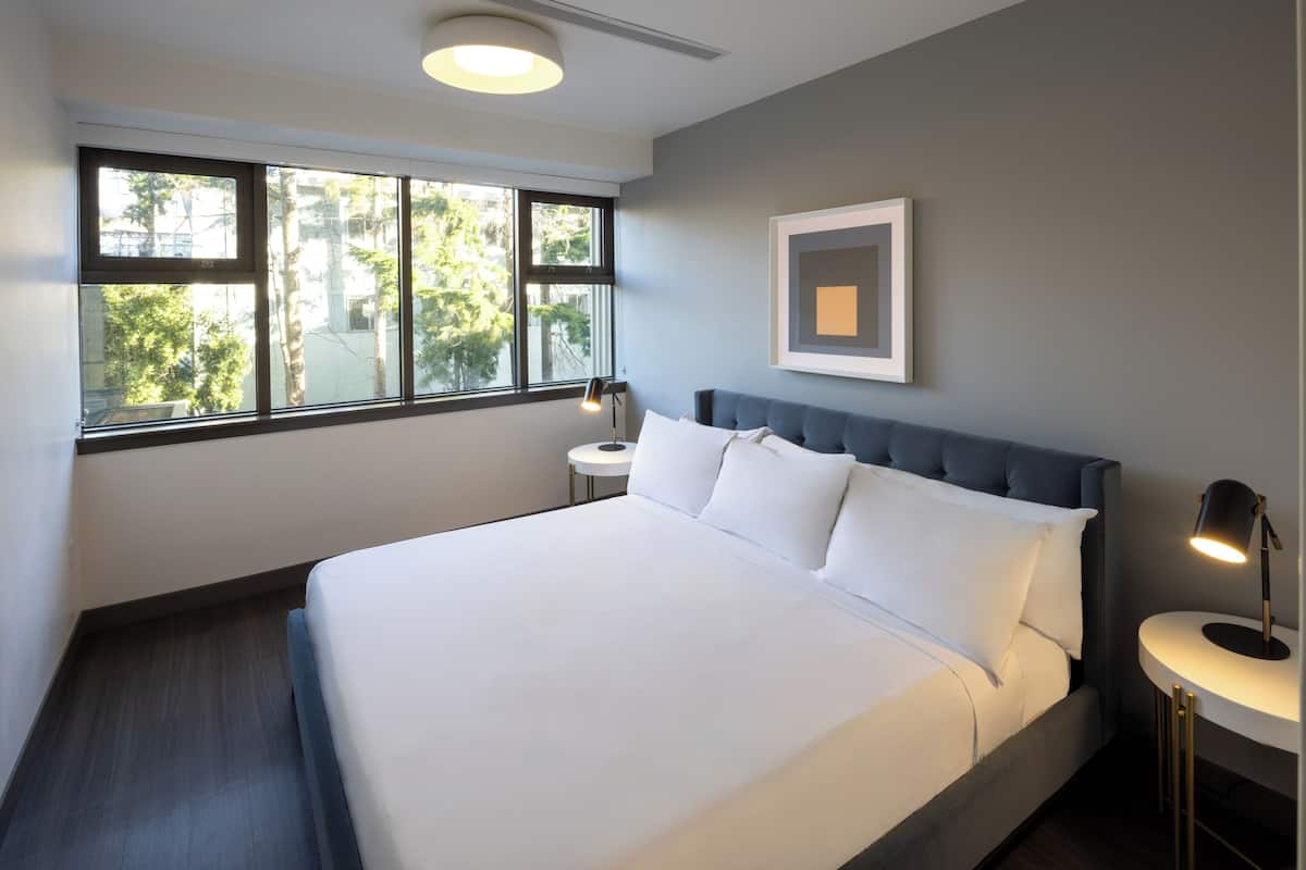 Una habitación de hotel moderna con una cama grande, una mesita de noche y una ventana con vistas a árboles.