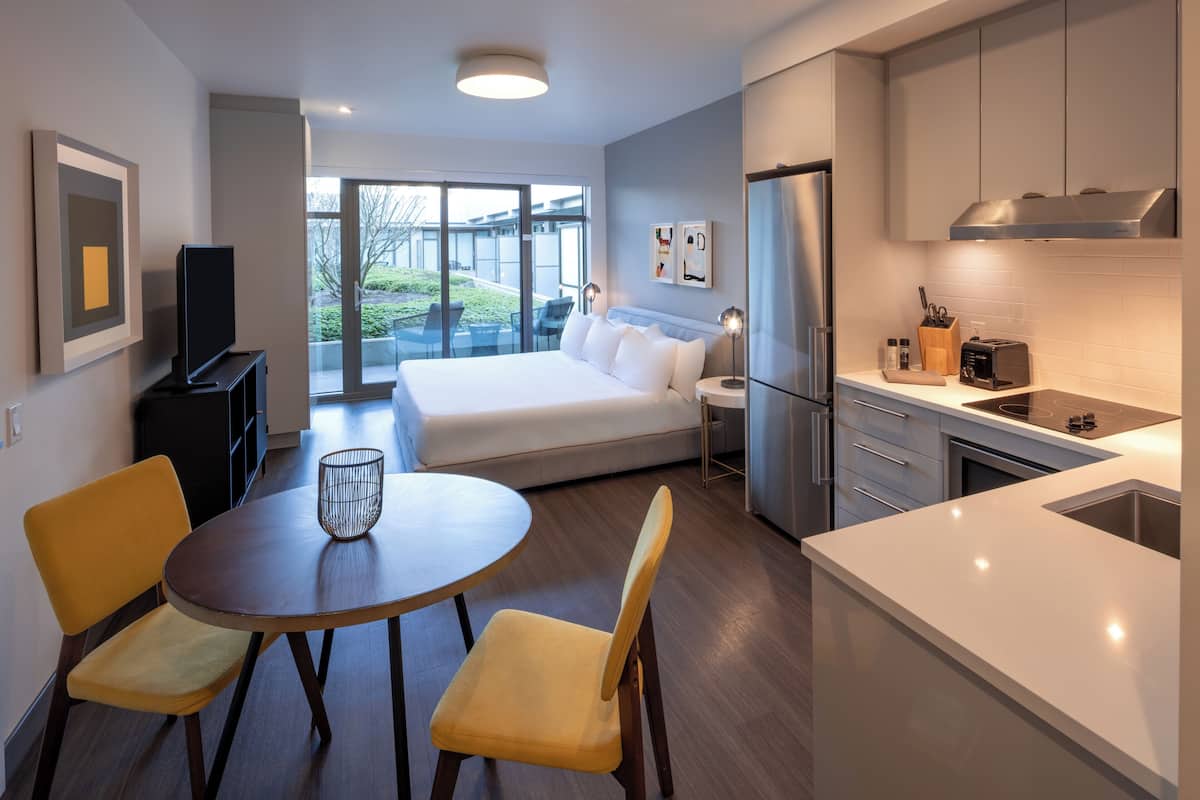 Una habitación de hotel moderna con cama, comedor con mesa y sillas, cocina con refrigerador y estufa, y una puerta corrediza de vidrio al exterior.