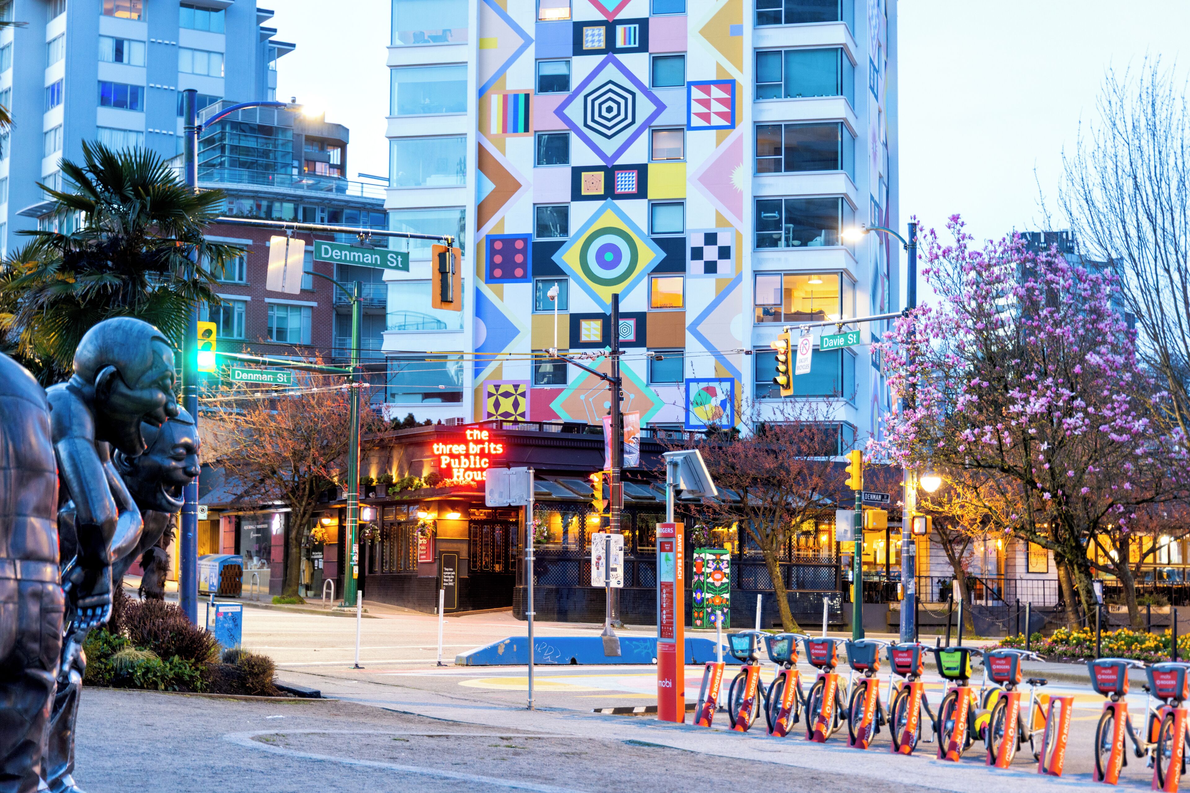 A building with a colorful geometric facade, a street with a bike-sharing station, and a statue of a man.