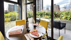 A dining table with a pizza box, a plate with pizza, a wine bottle, and glasses, set in a room with large windows overlooking a cityscape. - Revival English Bay, Vancouver  (Vancouver)
