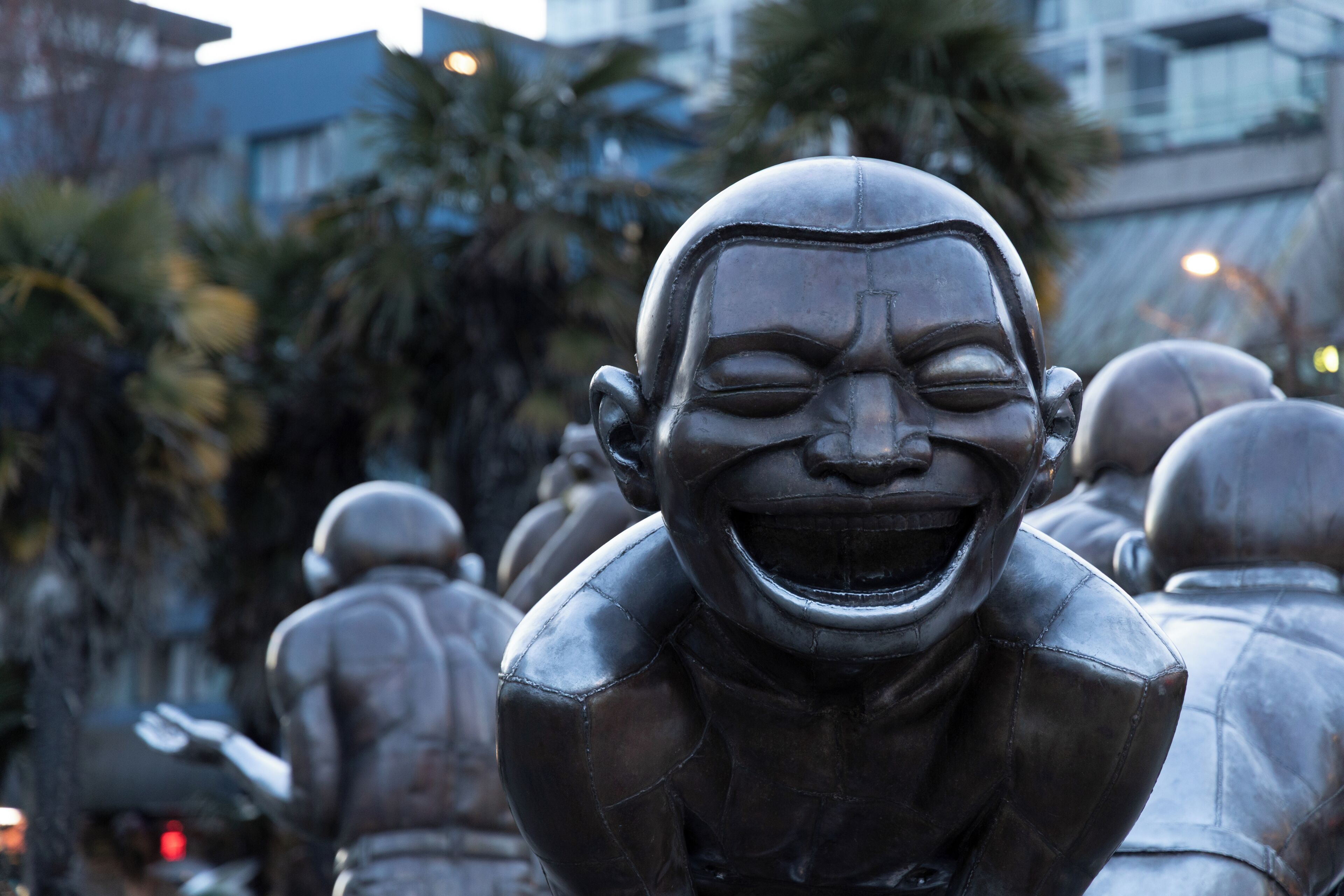 Uma estátua de um homem com um sorriso largo, vestindo terno, em ambiente externo com outras estátuas semelhantes e edifícios ao fundo.