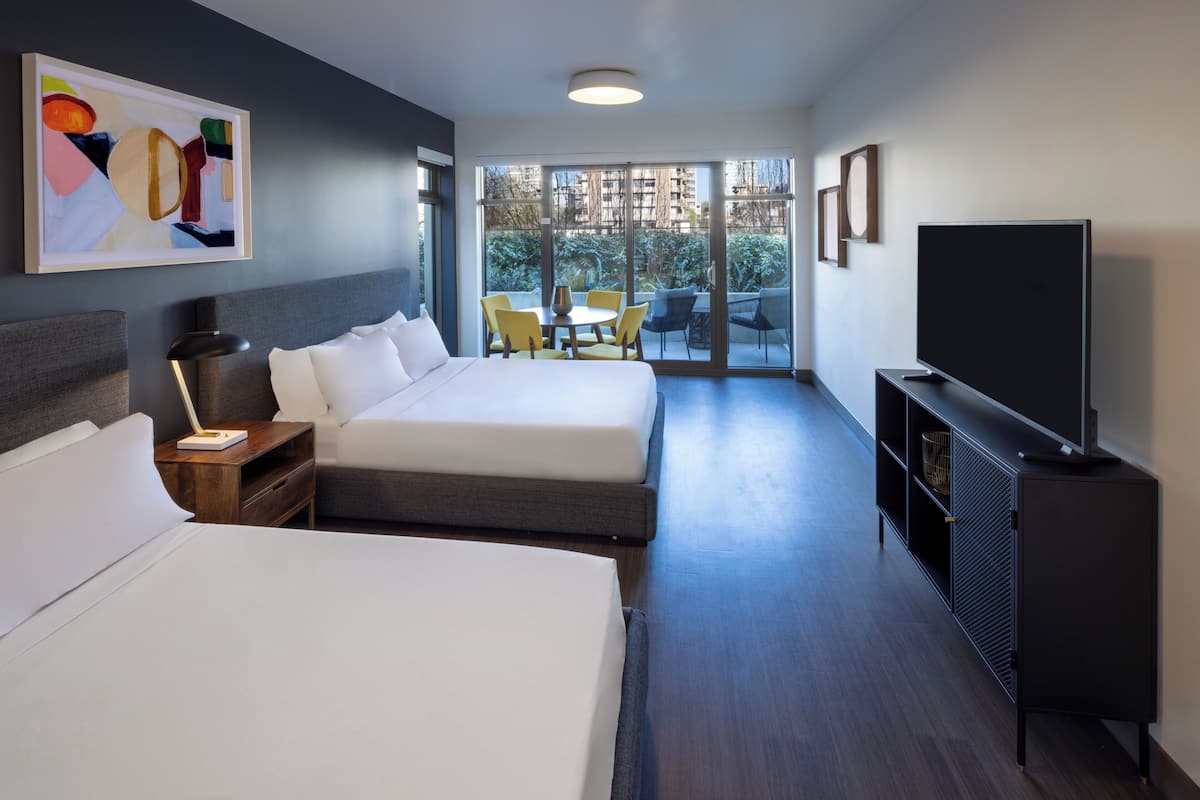 Habitación de hotel con dos camas, televisión, escritorio y balcón con mobiliario exterior.