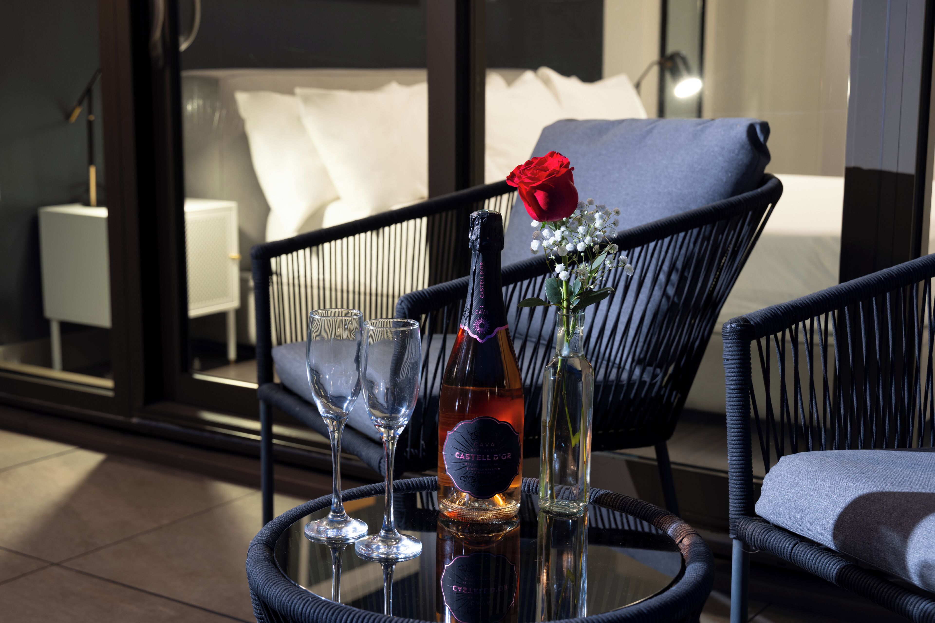 A table with a bottle, glasses, a rose, and a chair in a modern room with a view.
