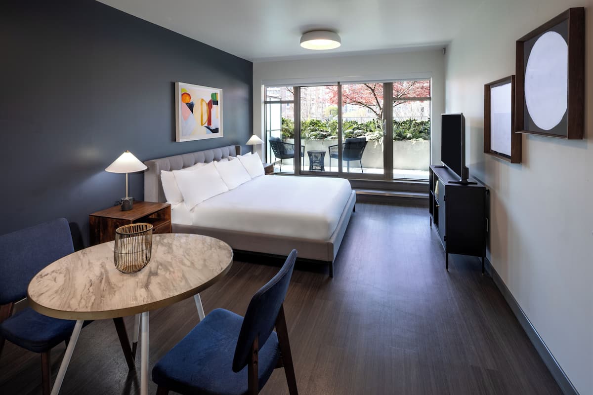 Habitación de hotel moderna con cama, una mesa redonda, dos sillas, televisor y ventana con vistas al exterior.