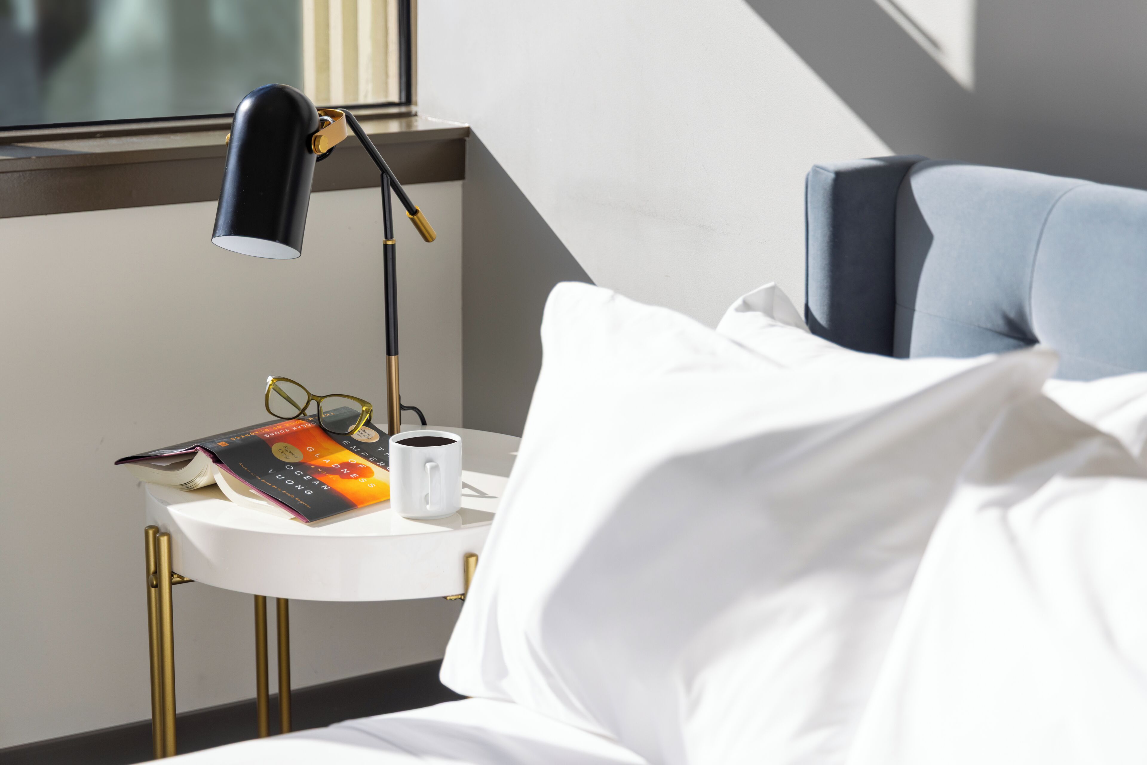 Een keurig opgemaakt bed met witte lakens, een nachtkastje met een lamp, een boek en een mok.