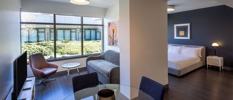 Una habitación de hotel moderna con una mesa de comedor de vidrio, asientos cómodos, una cama y vistas al exterior.