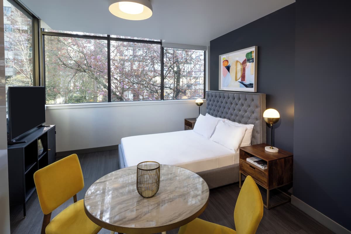 Habitación de hotel con cama, una mesa redonda con dos sillas, televisor y ventana con vista a árboles.
