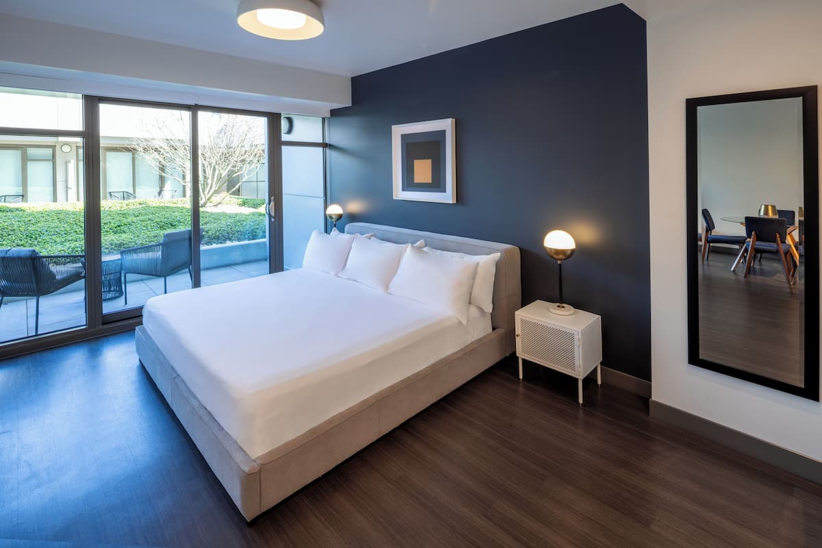 Un dormitorio moderno con una cama grande, una mesita de noche pequeña, un cuadro en la pared y un ventanal con vistas al exterior.