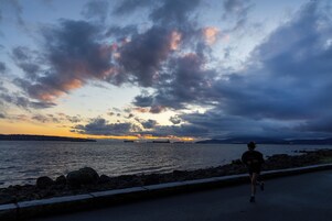 Una carretera costera con un corredor, un litoral rocoso y un atardecer sobre el agua.