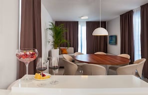 A modern hotel room with a dining area featuring a round wooden table, beige chairs, and a white tablecloth. There is a large potted plant, a sofa, and a hanging light fixture. - Luxury Seafront Villa Trogir 1 with 2 pools & Gym (Trogir)