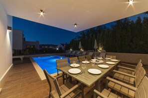 Uma área de refeições à beira da piscina, com uma mesa posta para seis pessoas, rodeada por espreguiçadeiras.