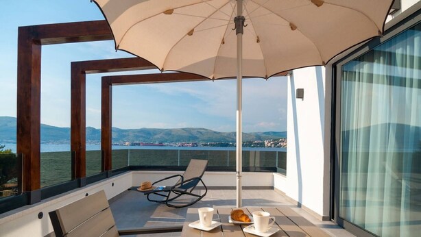 Ein Balkon mit Tisch, Stühlen und Sonnenschirm, mit Blick auf ein Gewässer und eine entfernte Landschaft.
