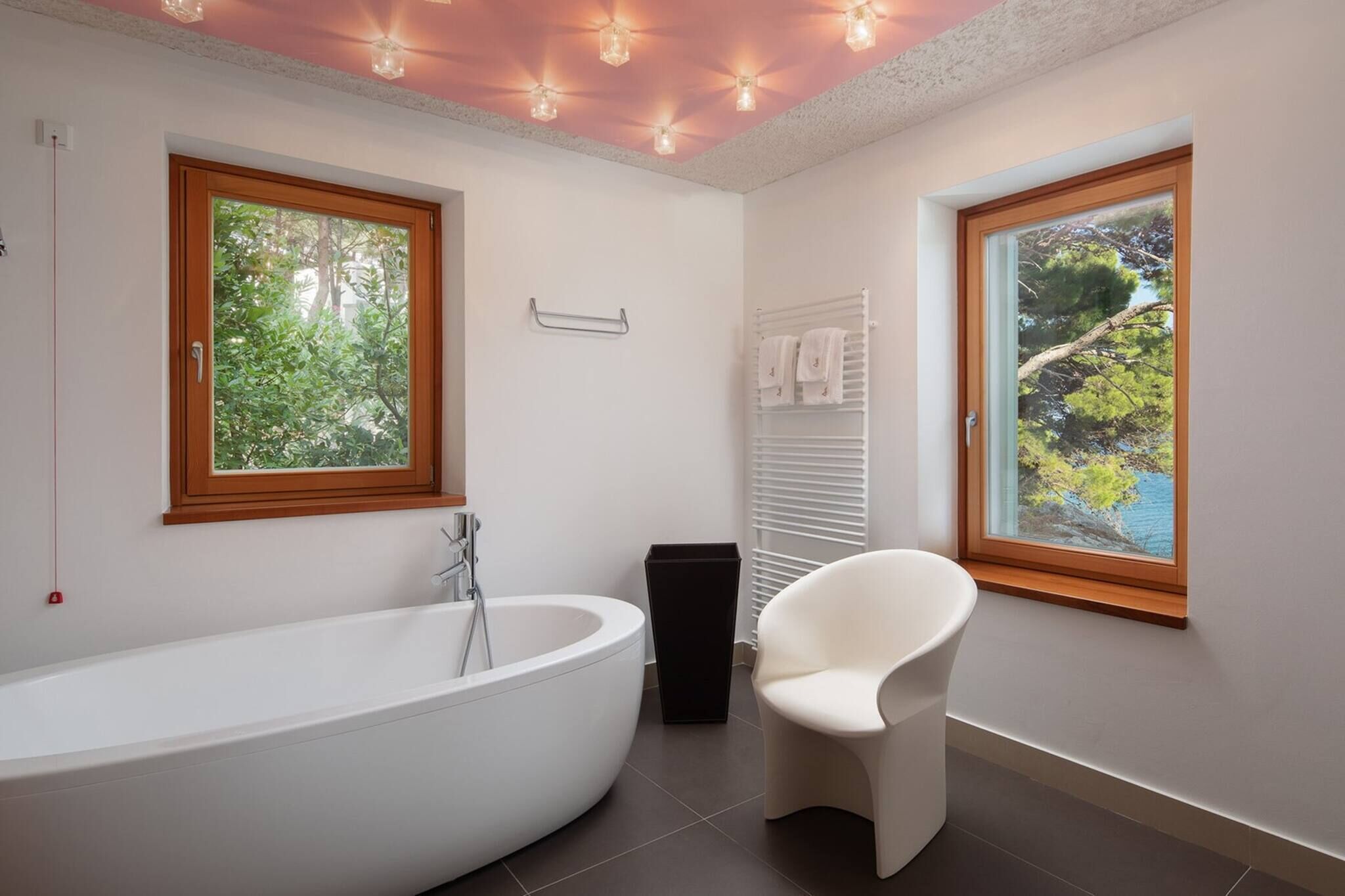 Een moderne badkamer met een vrijstaand bad, een raam met uitzicht op groen en een witte stoel.
