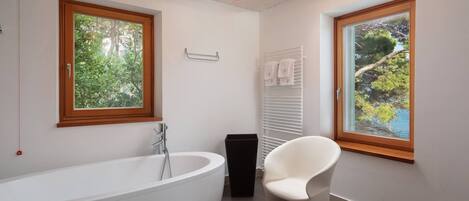 Een moderne badkamer met een vrijstaand bad, een raam met uitzicht op groen en een witte stoel.