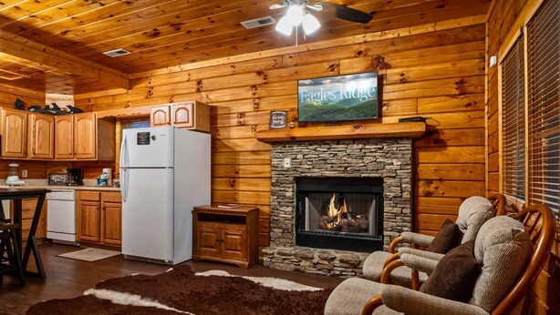 A cozy cabin interior with a stone fireplace, wooden walls, a kitchen area, and a seating area with a fur rug.