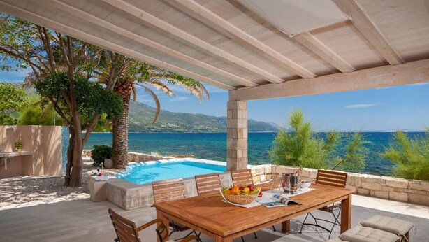 An outdoor dining area with a pool, wooden table, and chairs, overlooking a clear blue sea and mountains.
