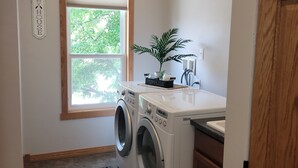 A laundry room with a washing machine, a sink, a window with blinds, and a sign on the wall. - Lakeview Loft (Kewaunee)
