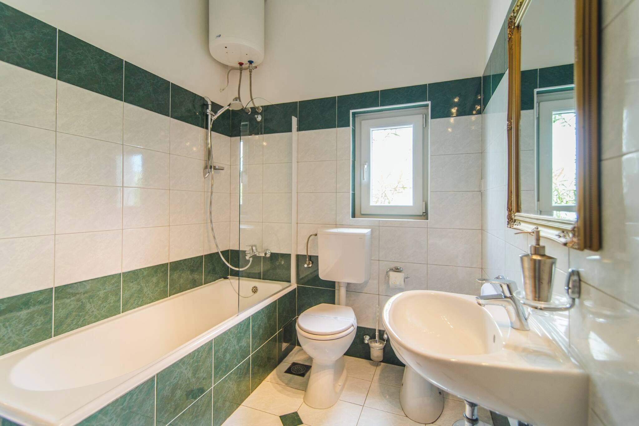 Een badkamer met bad, toilet en wastafel, met groene en witte tegels en een raam.