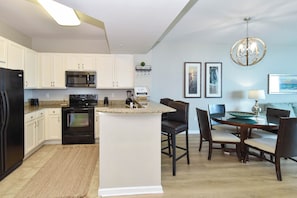Una cocina con armarios blancos, un refrigerador negro, un microondas y un horno. Hay un comedor con una mesa y sillas.