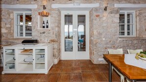Une pièce aux murs de pierre et au plafond en bois, un meuble blanc aux portes vitrées, une table recouverte d’une nappe blanche, et une vue à travers une porte en verre.