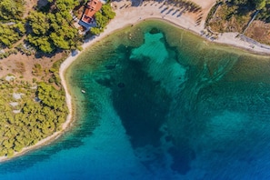 An aerial view of a coastal area with clear turquoise waters, a sandy beach, and dense greenery.