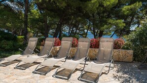 A row of sun loungers with metal frames and beige fabric, arranged on a stone patio with a backdrop of trees and flowering plants. - Luxury Villa Bol Oasis Brac With Heated Pool (Bol)