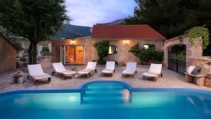 A poolside area with lounge chairs, a stone building with a red-tiled roof, and a view of the mountains.