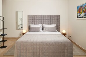 A bedroom with a tufted headboard, two bedside lamps, a bed with a quilted cover, a bedside table with a lamp, a mirror, and a framed artwork on the wall.