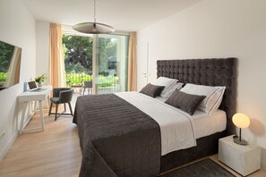 Une chambre avec un grand lit, un bureau et une chaise, un balcon avec vue sur les arbres et une lampe moderne sur la table de chevet.