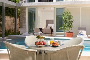 Un coin petit-déjeuner aménagé au bord de la piscine, avec des chaises en osier, un plateau de fruits et un verre de jus d’orange.