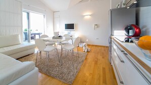 Un salon moderne avec un canapé blanc, une table à manger et des chaises, une cuisine équipée d’un réfrigérateur et d’une cuisinière, et un petit chien.