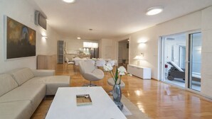 Un salon moderne avec un canapé, une table basse et des fauteuils, un coin repas avec une table et des chaises, et une cuisine en arrière-plan.