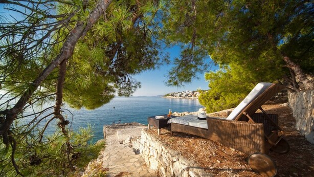 Une vue sur la mer avec un fauteuil de jardin en osier, un mur de pierre et une mer d’un bleu limpide.