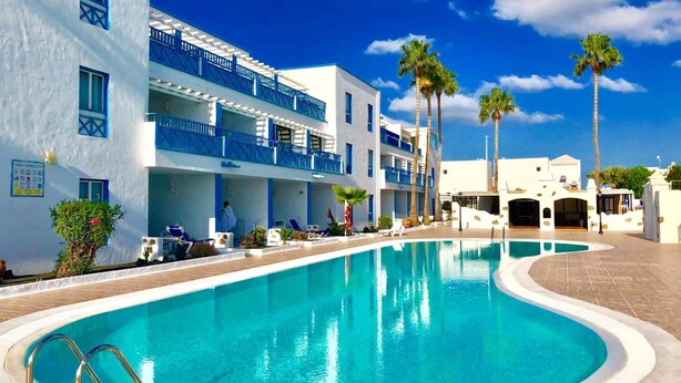 A poolside view of a hotel with a clear blue pool, white buildings with blue accents, and palm trees. - Aguazul (Tías)