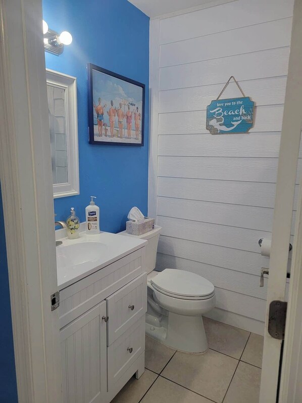 A bathroom with a blue wall, white vanity, and a framed picture above the sink. - Nauti Escape Across The Street From Beach Access (Madeira Beach)