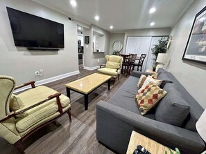 A living room with a flat-screen TV, a grey sofa, a yellow armchair, and a wooden coffee table. - Sunny Renovated 2BR Condo · Pool · Near DC (West Springfield)