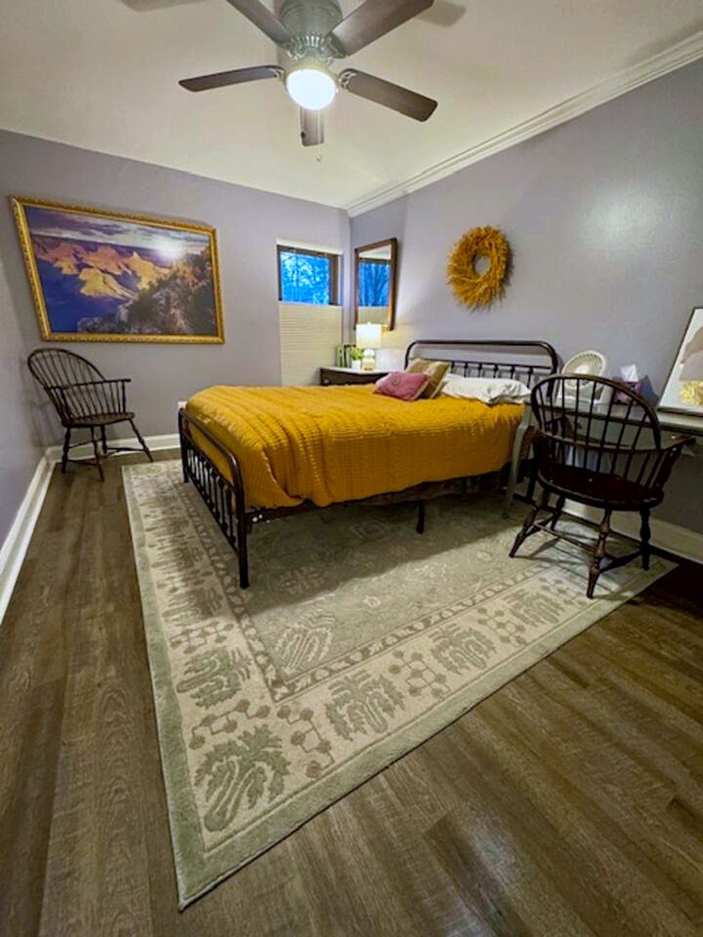 A bedroom with a bed, two chairs, a ceiling fan, a rug, a painting, and a wreath on the wall.