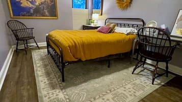 A bedroom with a bed, two chairs, a ceiling fan, a rug, a painting, and a wreath on the wall.