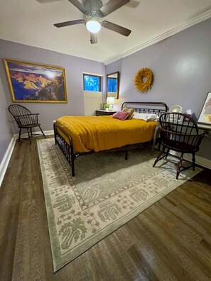A bedroom with a bed, two chairs, a ceiling fan, a rug, a painting, and a wreath on the wall. - Sunny Renovated 2BR Condo · Pool · Near DC (West Springfield)