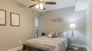 A bedroom with a bed, a ceiling fan, a nightstand with a lamp, and wall art.