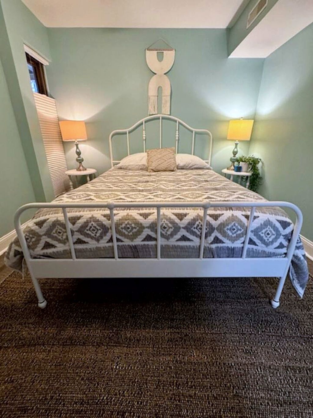 A bedroom with a bed, two bedside tables, a wall-mounted lamp, and a window with blinds.