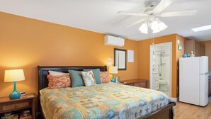Un dormitorio con cama, mesitas de noche, ventilador de techo, refrigerador blanco y baño con ducha e inodoro.