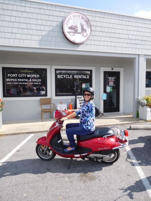 A person riding a red scooter in front of a building with signage for 'Port City Moped' and 'Bicycle Rentals'.