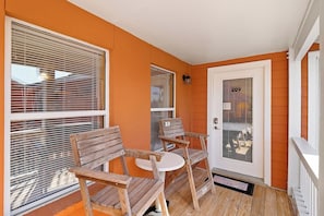 A porch with two wooden chairs and a small round table, a door with the number 209, and a window with blinds.