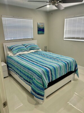 A bedroom with a bed, white nightstands, a ceiling fan, and windows with blinds.