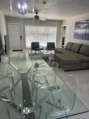 A modern living room with a glass dining table, two chairs, a sofa, and a ceiling fan.