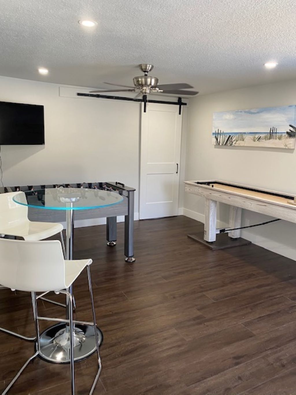 Een moderne kamer met een ronde glazen tafel, witte stoelen, een plafondventilator, een tv aan de muur en een schilderij.